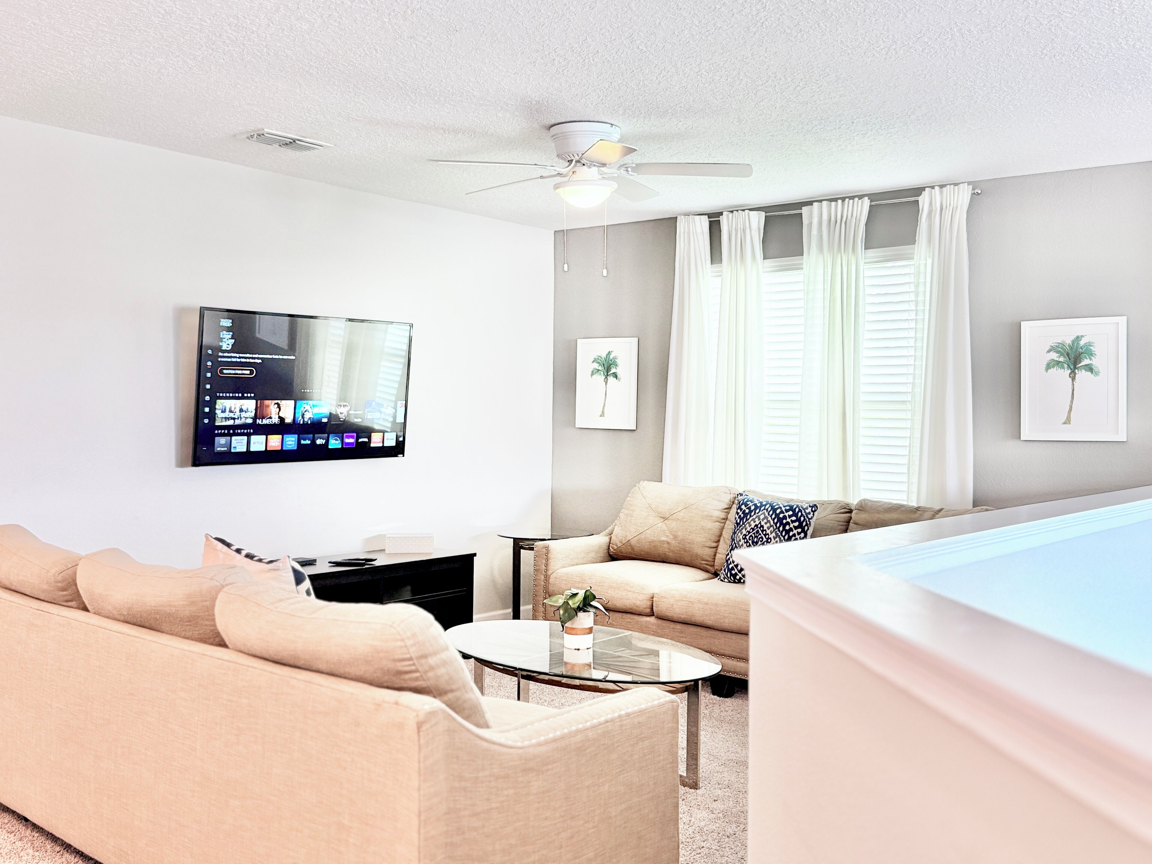 Ein Wohnzimmer mit Flachbildfernseher, Deckenventilator, Sofa, Couchtisch und einem Fenster mit Vorhängen.