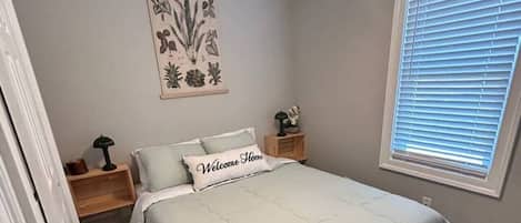 Un dormitorio con cama, ventilador de techo, ventana con persianas, mesita de noche y una obra de arte colgada en la pared.
