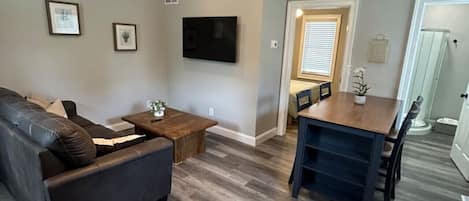 Una sala de estar con ventilador de techo, televisor de pantalla plana, sofá oscuro, una mesa de centro de madera y una zona de comedor con mesa y sillas.