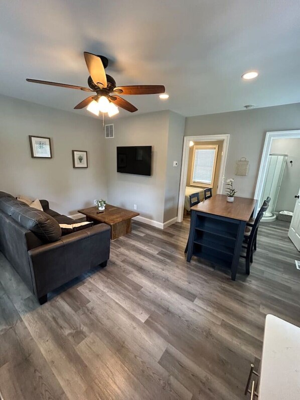 A living room with a ceiling fan, a flat-screen TV, a dark sofa, a wooden coffee table, and a dining area with a table and chairs. - Whispering Pines Cottage (Egg Harbor City)