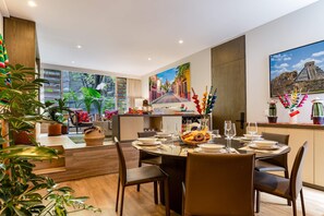 A dining area with a table set for two, a painting of a street scene, and a view of a pyramid through a window. - Colorful Roma Norte 3BR/3BA, 2 patios, AC (Mexico City)