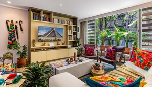 A living room with a large TV, bookshelves, a sofa, and a view of trees through large windows. - Colorful Roma Norte 3BR/3BA, 2 patios, AC (Mexico City)