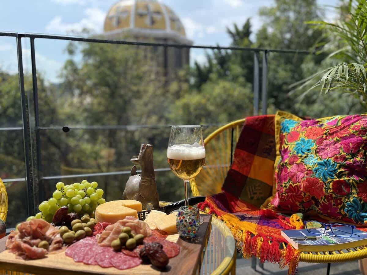 Un balcón con una mesa dispuesta con comida y bebidas, una silla de mimbre y una manta colorida.