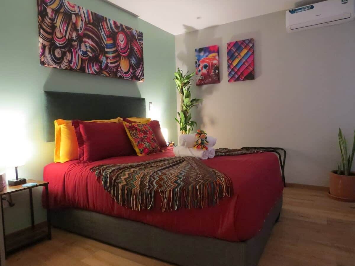 A bedroom with a bed, pillows, a blanket, a nightstand, a potted plant, and artwork on the walls.