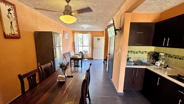 Ein Essbereich mit Holztisch und -stühlen, einem Deckenventilator und einer Küche mit dunklen Schränken und einer gefliesten Spritzschutzwand.