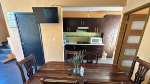 Ein Essbereich mit Holztisch und Stühlen, einem an der Wand montierten Flachbildfernseher und einer Küchenzeile mit Schränken und einer Mikrowelle.