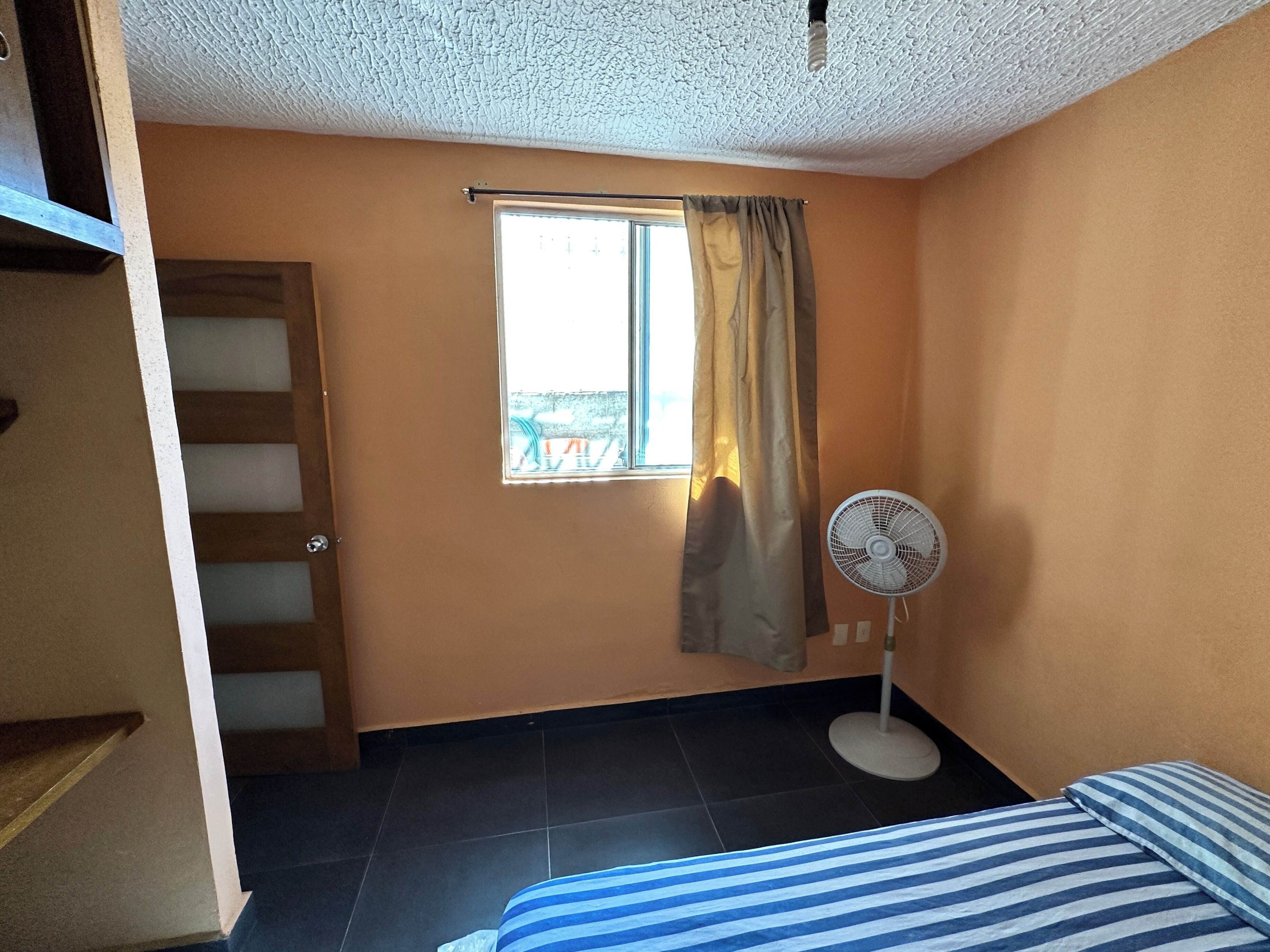 A room with a bed, a standing fan, a window with curtains, and a door leading to a staircase.