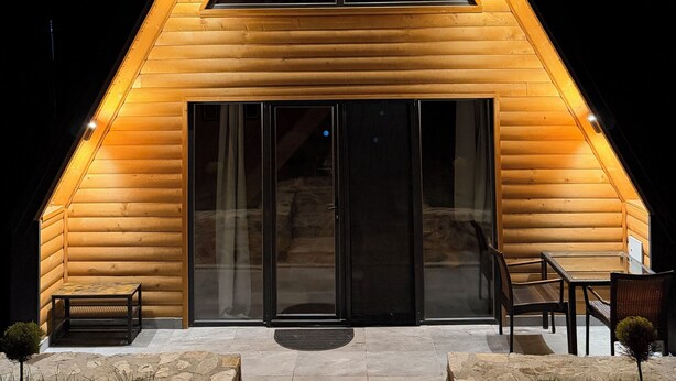 Une cabane triangulaire en bois, dotée d’une grande porte-fenêtre, d’un escalier en pierre et d’un espace salon extérieur.