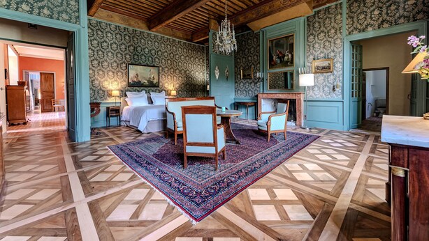 Een kamer met een groot bed, twee stoelen, een tafel, een kroonluchter en behangpapier met een patroon.
