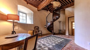 A dining area with a wooden table, chairs, a patterned rug, a lamp, and a spiral staircase. - Tour Ouest Vacation Rental - Château de Vachères (Montclar-sur-Gervanne)