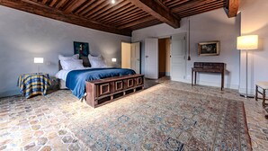 Um quarto com uma cama, um tapete estampado, um banco de madeira, uma mesinha de cabeceira, um abajur, um quadro e uma porta.