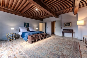 A bedroom with a bed, a patterned rug, a wooden bench, a nightstand, a lamp, a painting, and a door.