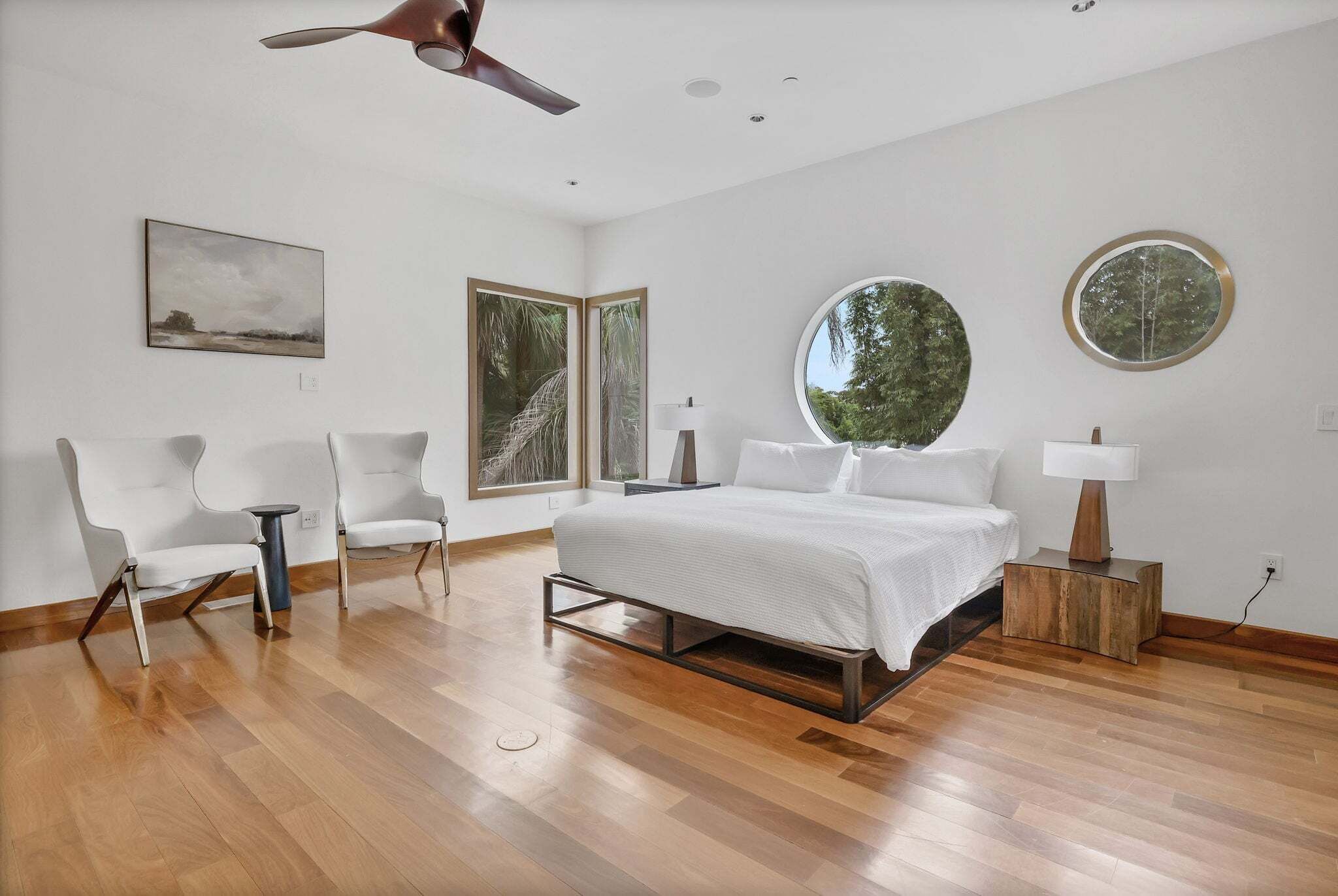 Une chambre moderne avec un grand lit, deux fauteuils, un miroir rond et un ventilateur de plafond.