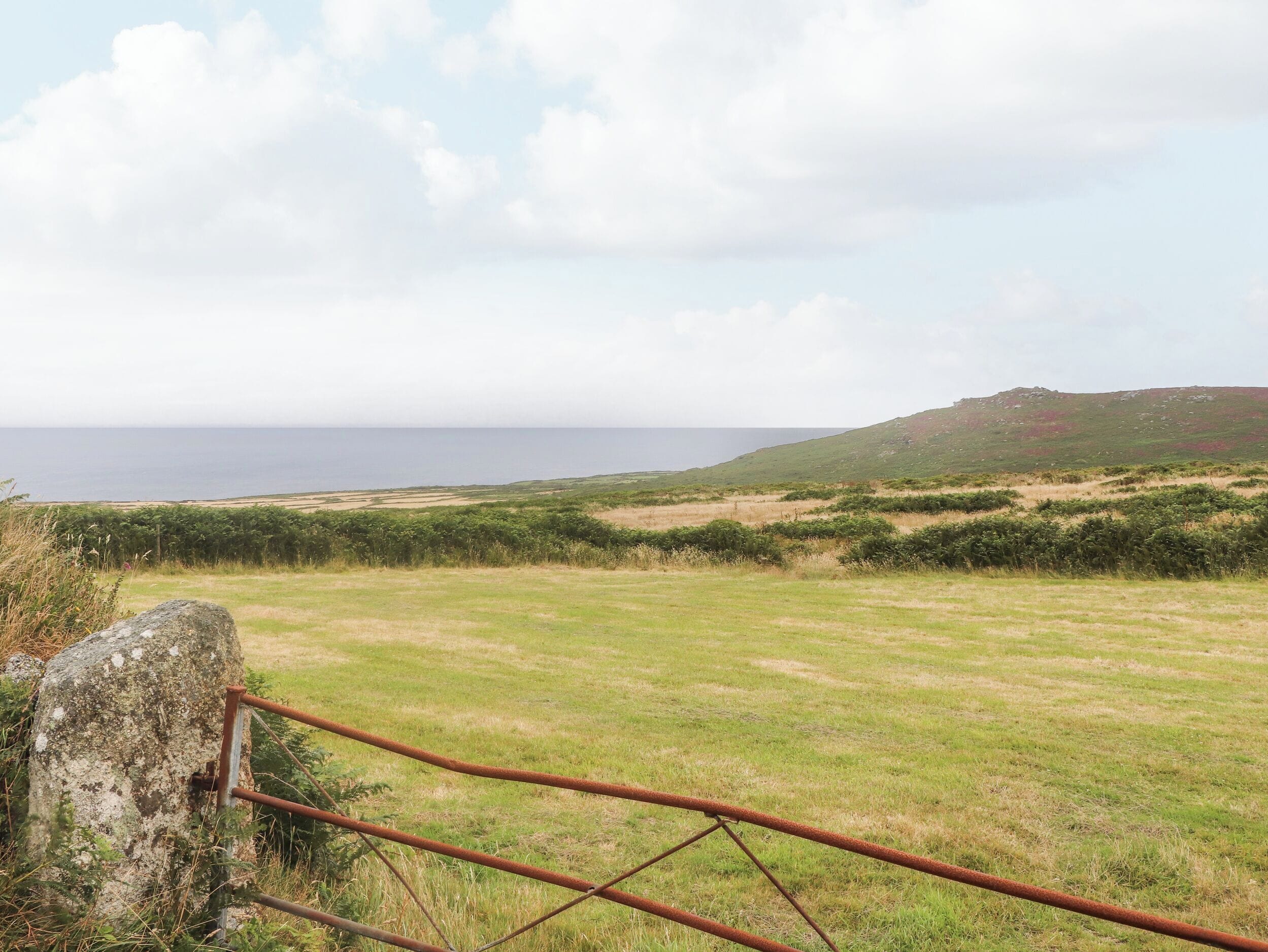 Paisatge costaner amb un camp herbaci, una reixa metàl·lica i una vista llunyana del mar i les muntanyes.