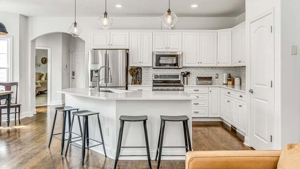 Una cocina moderna con gabinetes blancos, una isla central con taburetes, electrodomésticos de acero inoxidable e iluminación colgante.