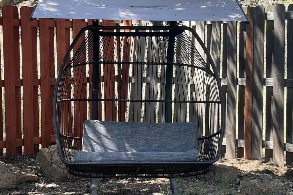 Ein Hängekorbstuhl mit Metallgestell und Kissen, draußen aufgestellt, mit einem Holzzaun im Hintergrund.