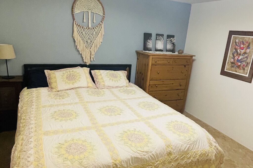 A bed with a floral bedspread, a wooden dresser, a macrame wall hanging, and a framed picture on the wall.