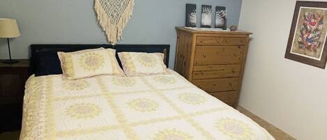A bed with a floral bedspread, a wooden dresser, a macrame wall hanging, and a framed picture on the wall.