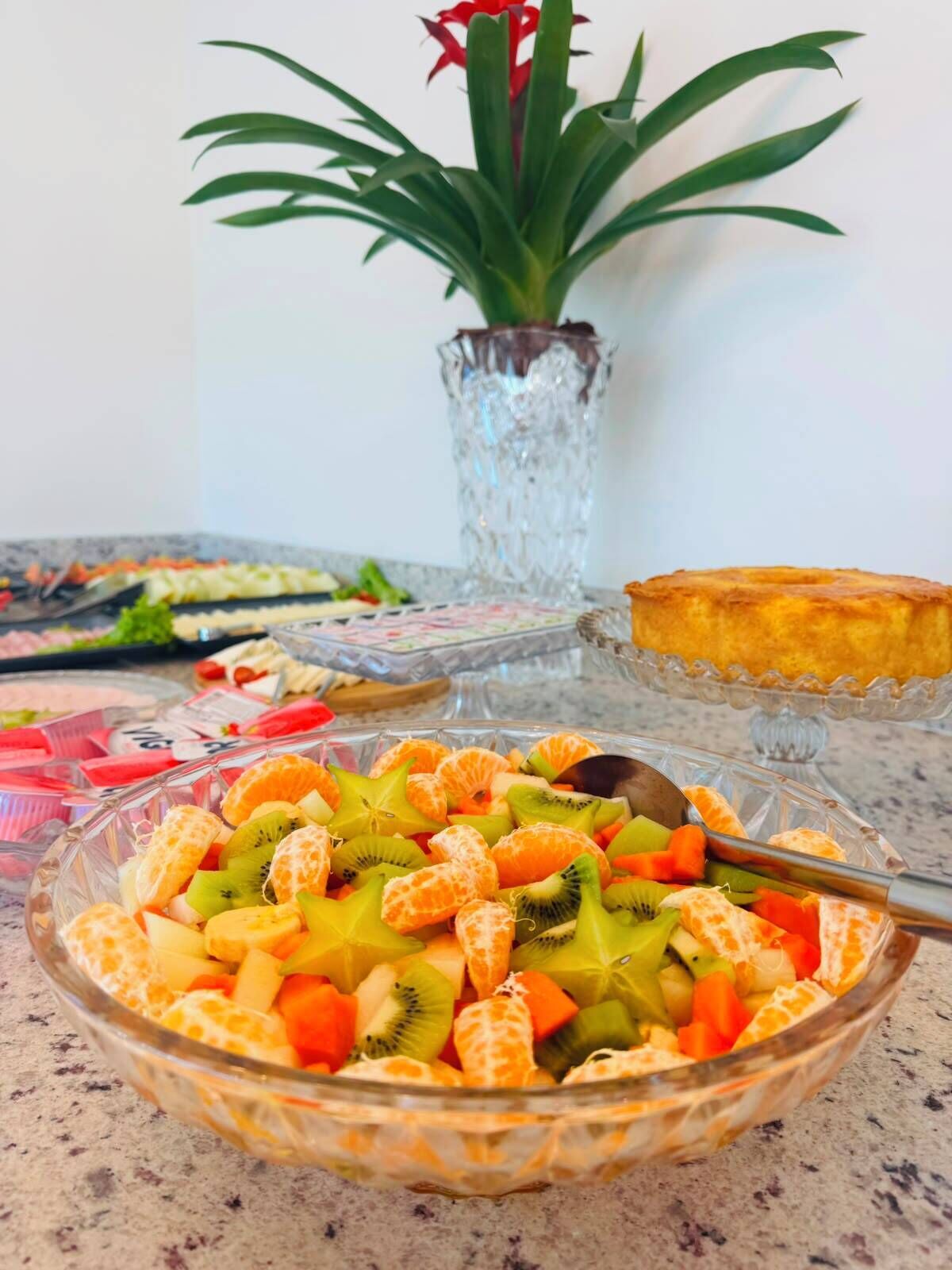 Un tazón de ensalada de frutas sobre una mesa, con un florero de vidrio y una planta al fondo.