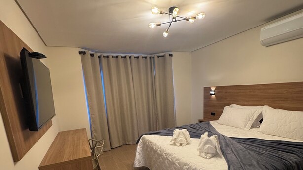 A hotel room with a bed, a TV mounted on the wall, a wooden bench, and a ceiling light fixture. - Le Bonne Ville Hotel (Campos do Jordão)
