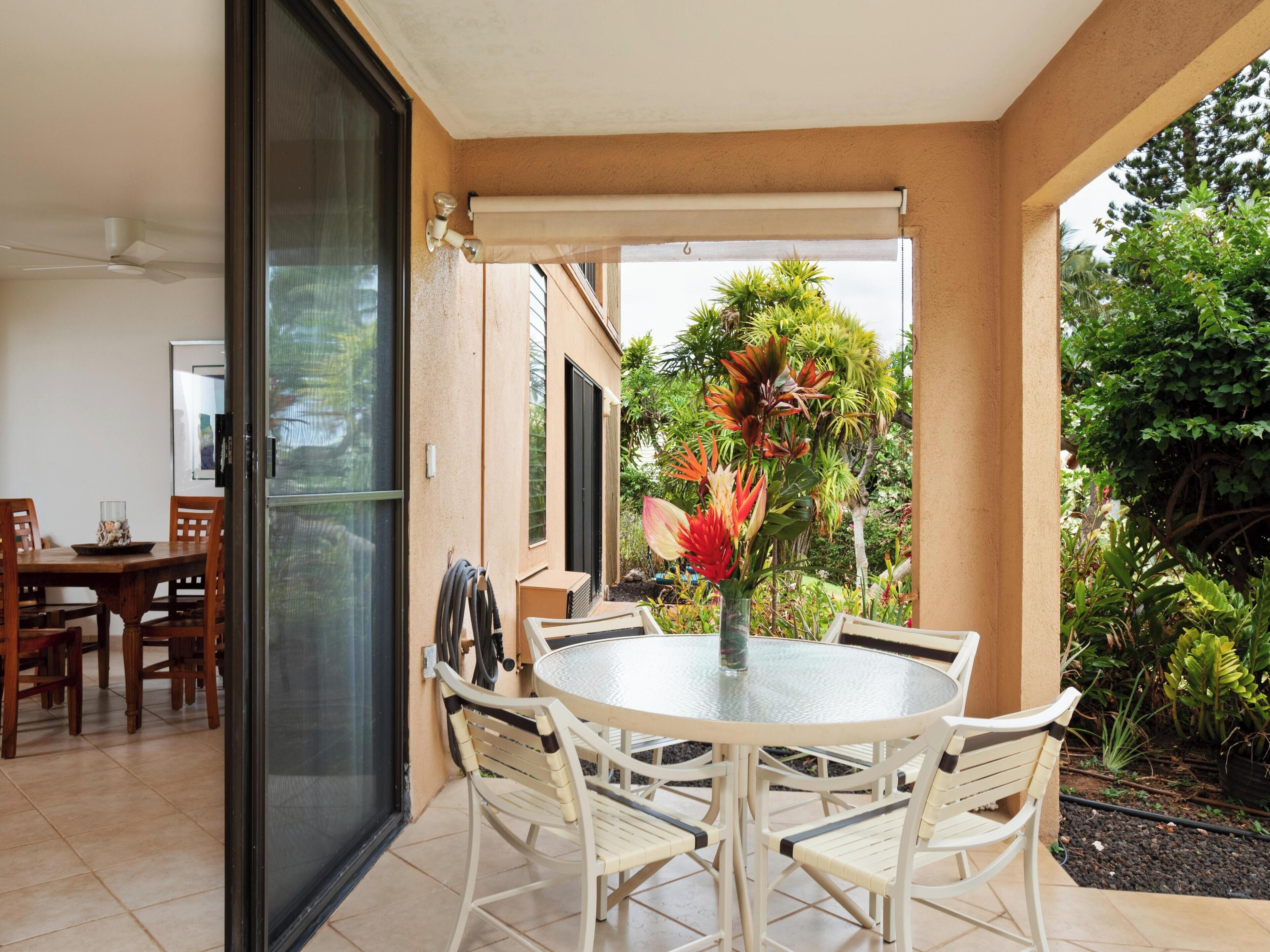 A patio with a round table and chairs, a glass door, and a view of a garden with tropical plants.