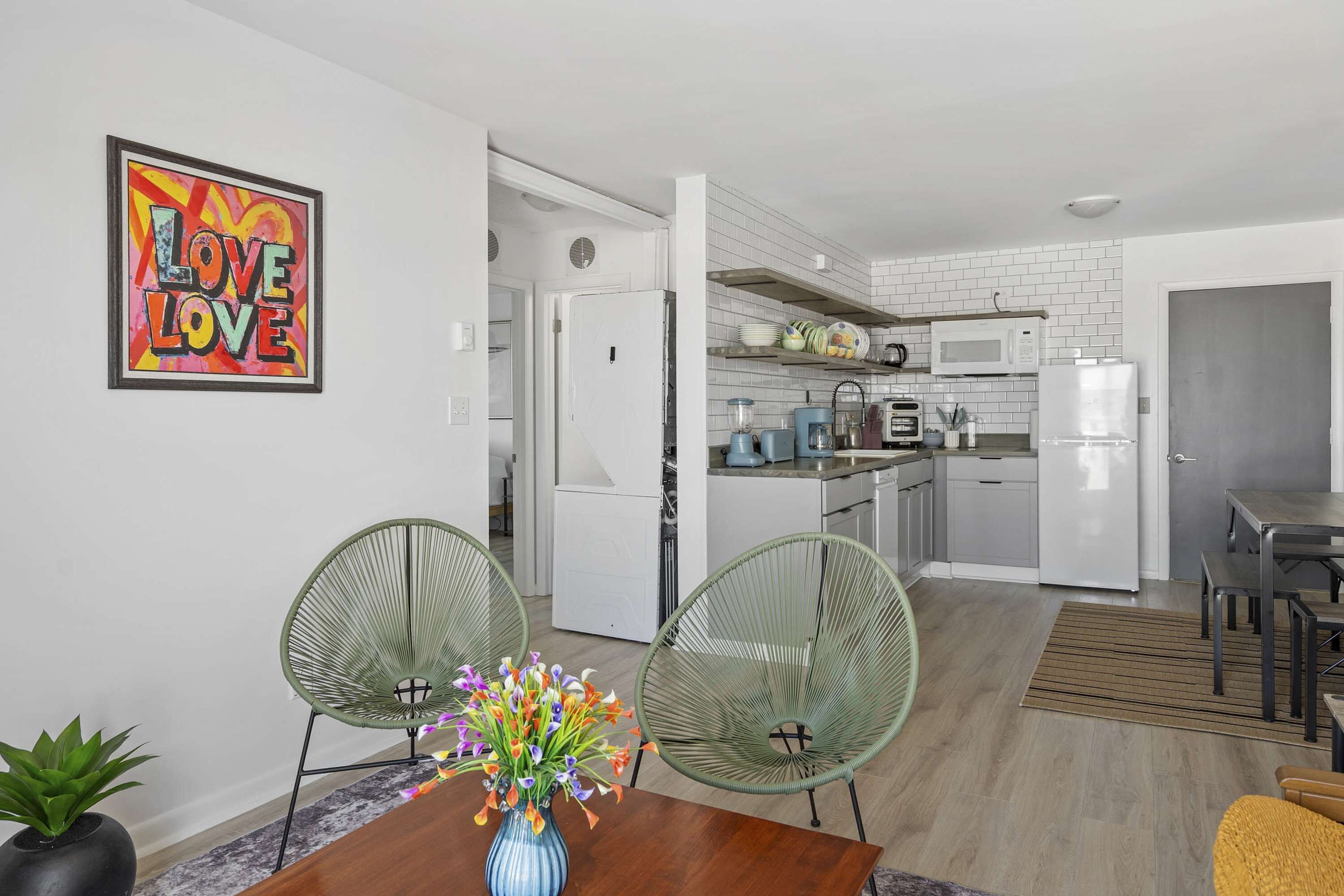 Een moderne keuken met witte apparaten, een eethoek met twee groene stoelen en een kunstwerk met de tekst 'LOVE LOVE' op de muur.