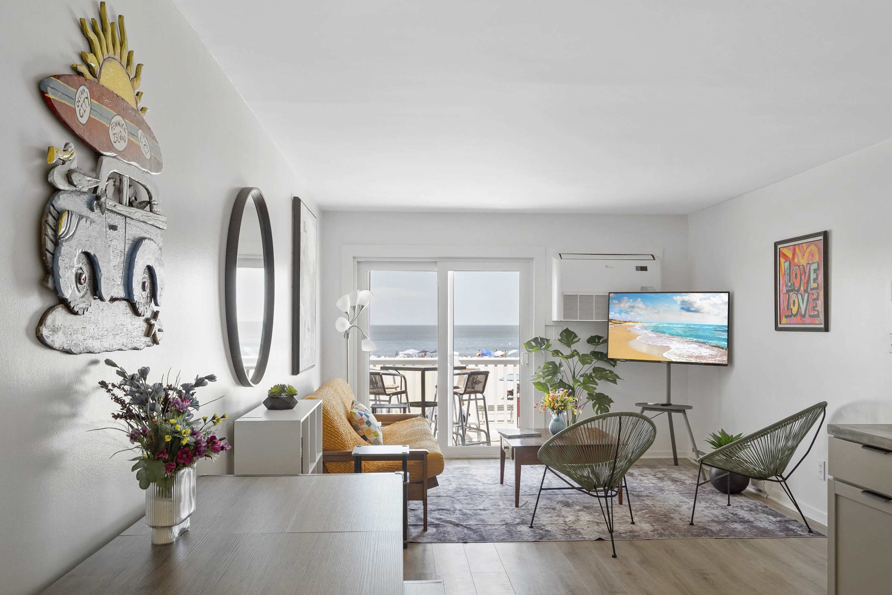 Ein modernes Wohnzimmer mit einer Couch, einem Esstisch mit Stühlen, einem Fernseher und einem Balkon mit Blick auf den Strand.