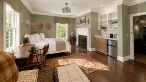 Une chambre avec un grand lit, un bureau et une chaise, une cheminée, ainsi qu’un coin cuisine équipé de placards et d’un évier.