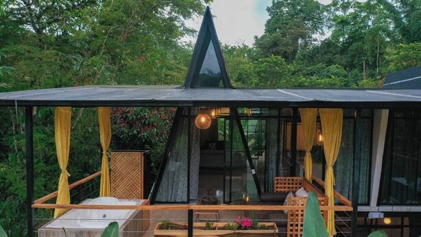 Una estructura moderna, con paredes de vidrio, terraza de madera, zona de estar y vistas a vegetación exuberante.