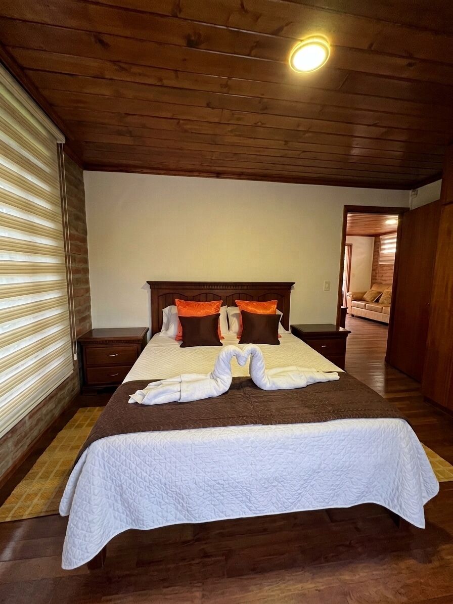 A hotel room with a wooden ceiling, a bed with white bedding, brown headboard, and orange pillows, a wooden nightstand, and a window with blinds.
