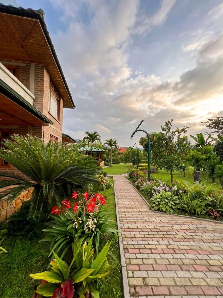 A pathway leading to a building with a brick facade, surrounded by lush greenery and a clear sky.