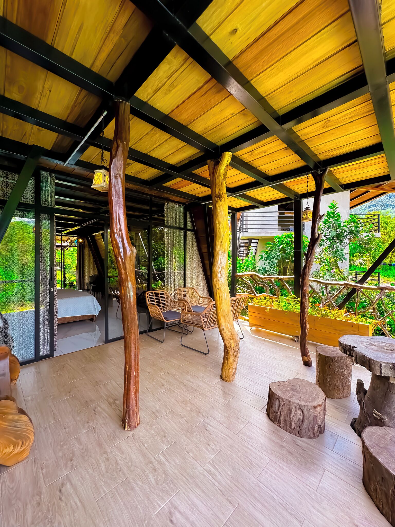 Un espacio al aire libre techado con suelo de madera, pilares rústicos de madera, mobiliario de mimbre y vistas al verdor.