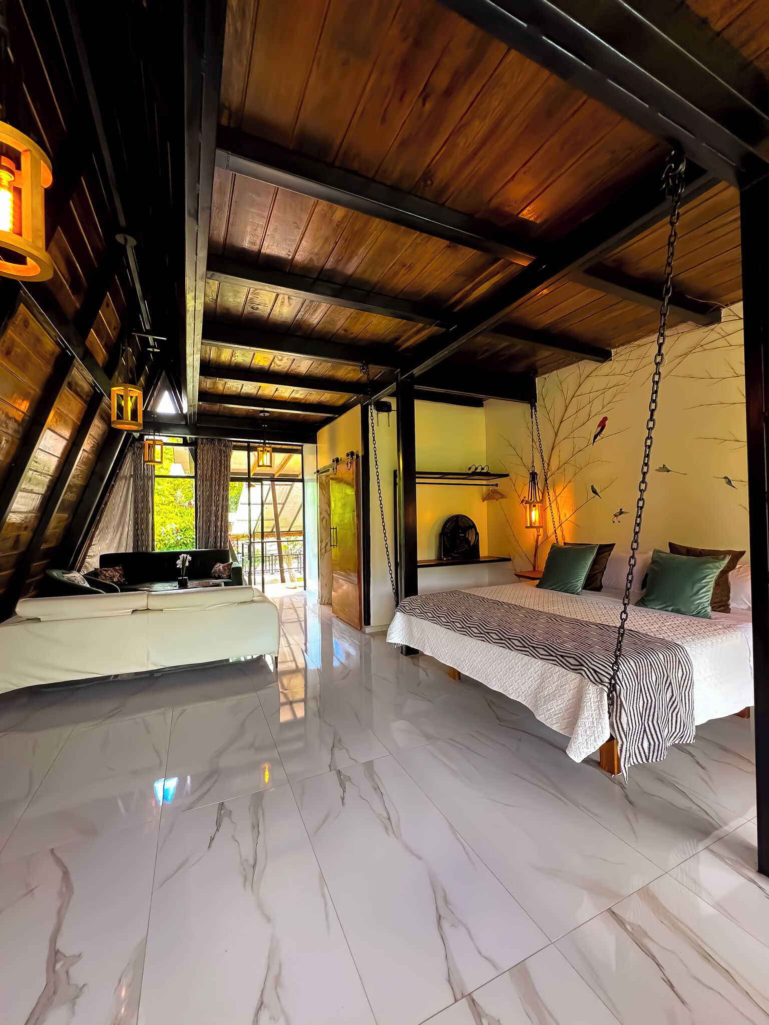 A spacious attic bedroom with a high ceiling, a bed with a patterned bedspread, a hanging light fixture, and a mural of birds on the wall.