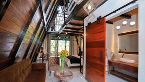 Una habitación con techo inclinado, un banco de madera, un sofá, una mesa con un jarrón de flores, una cama, un armario de madera y un baño con lavamanos y espejo.
