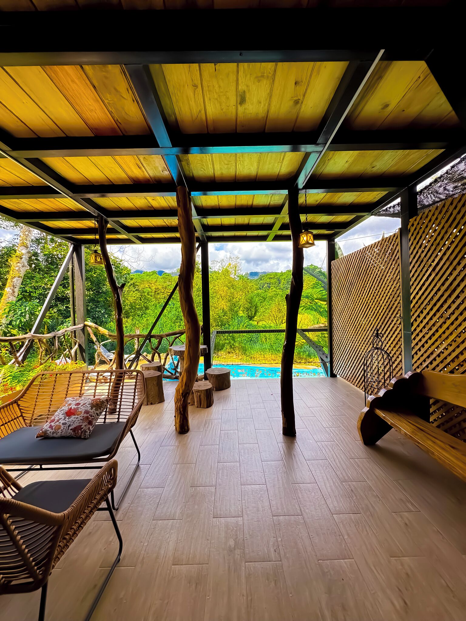 Une terrasse extérieure couverte, dotée d’un plancher en bois, de meubles en osier et offrant une vue sur une piscine.