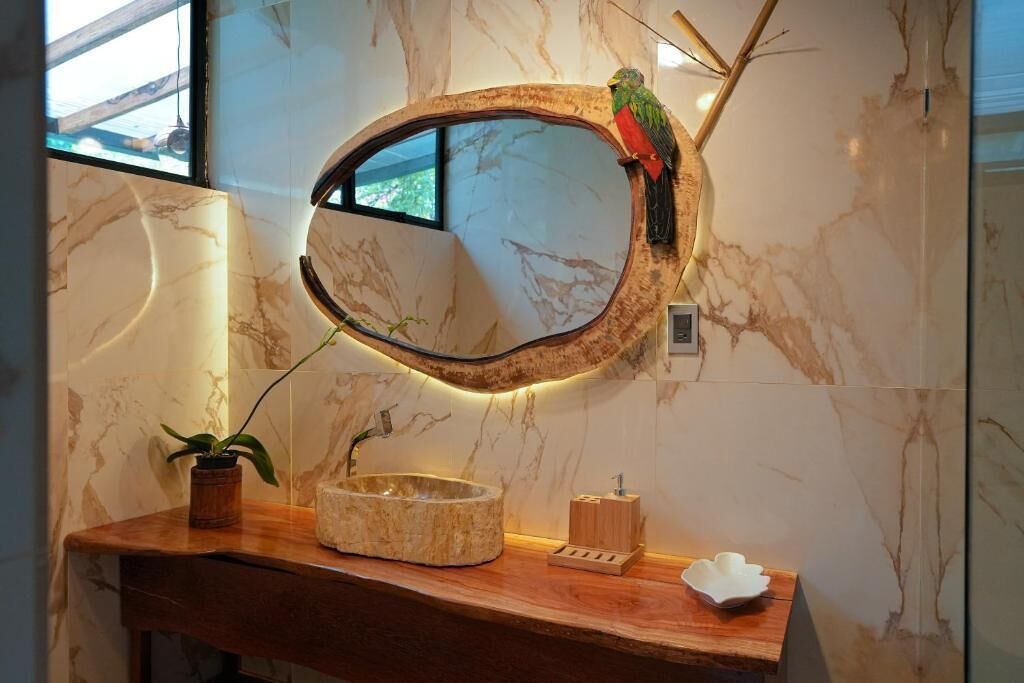A bathroom with a round mirror, a bird perched on it, a potted plant, a wooden vanity, and a marble wall.