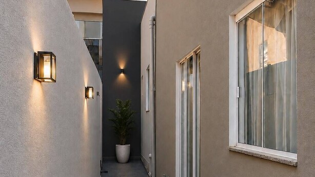 Un estrecho pasillo exterior con una planta en maceta, una puerta corredera de vidrio y luminarias fijadas a la pared.