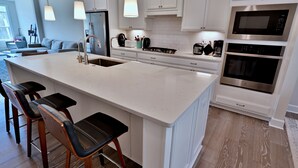 A modern kitchen with white cabinetry, a central island with bar stools, stainless steel appliances, and pendant lighting. - New Coastal Club Luxury Villa w Resort style Amenities | Sleeps 8 | 3BR / 2.5 Baths (Lewes)