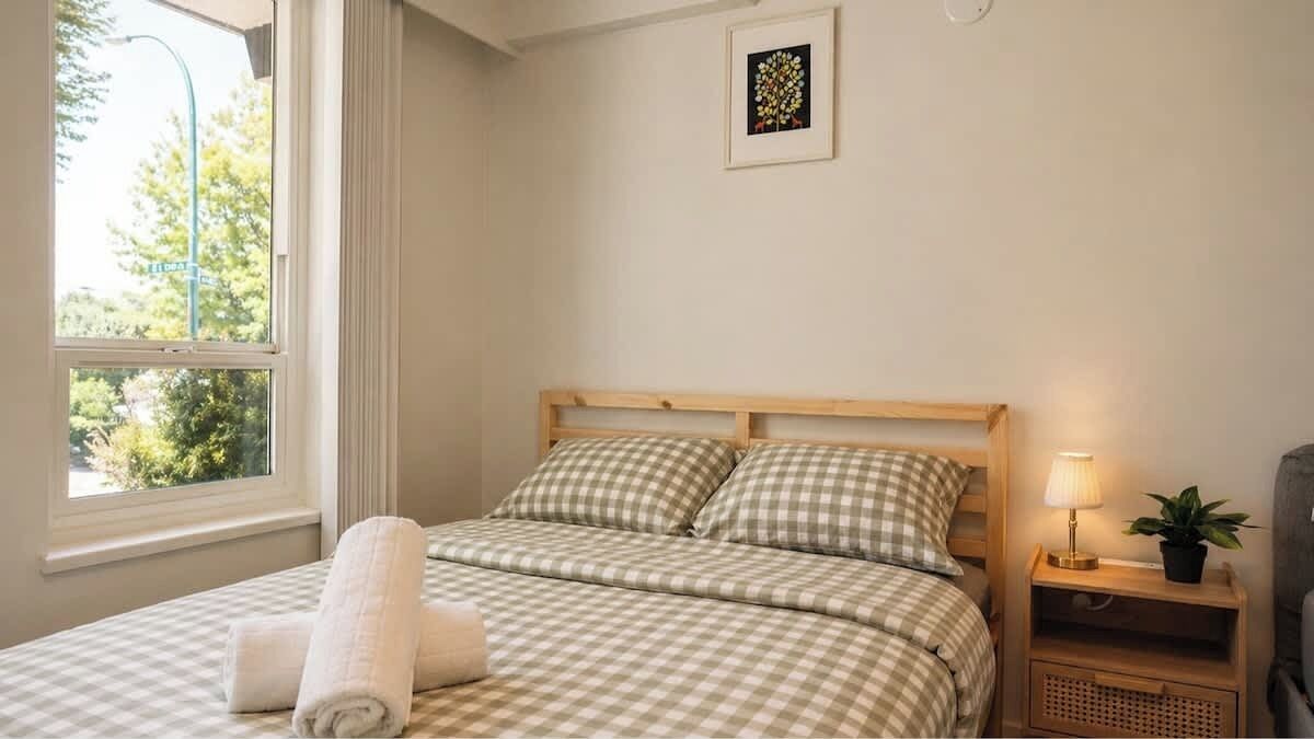 Una cama bien hecha con ropa de cama a cuadros, una lámpara de noche, una planta pequeña y un cuadro en la pared.