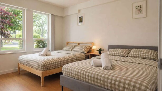 A hotel room with two beds, a window with a view of trees, a bedside table with a lamp, and a framed picture on the wall. - Comfy 3 Beds | 5-min Walk to SkyTrain | 20-min DT (Vancouver)