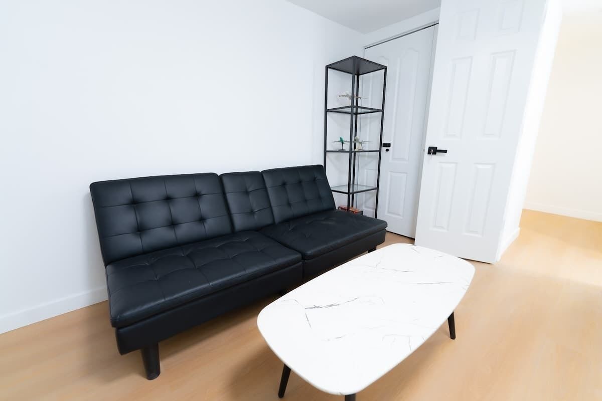 A black sofa, a white coffee table, and a glass shelving unit in a room with a white door and light-colored flooring.