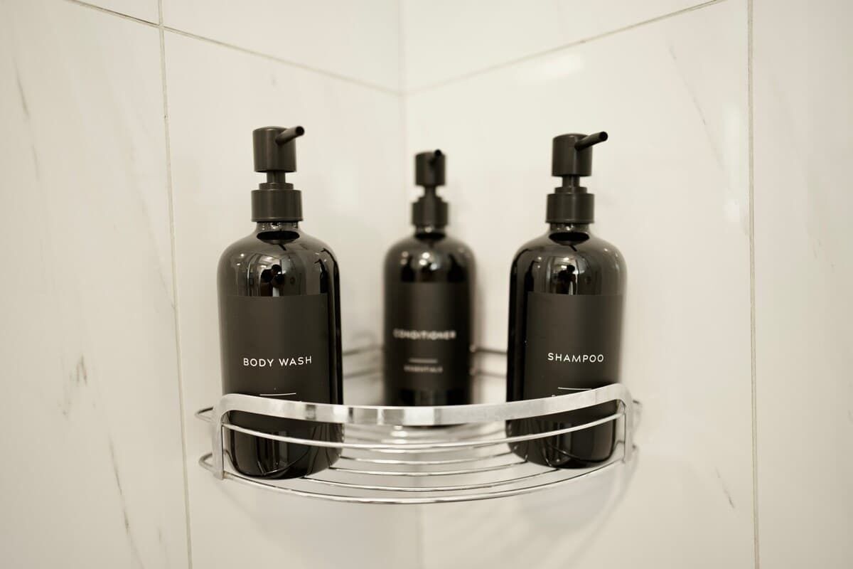 Un baño con pared de azulejos blancos, un estante de metal con tres botellas etiquetadas como 'BODY WASH', 'SHAMPOO' y 'SHAMPOO'.