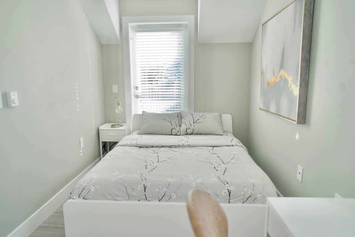 A bedroom with a bed, a nightstand, a window with blinds, and a wall-mounted picture.