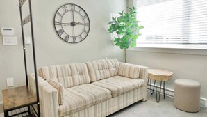 A modern living room with a striped sofa, a round clock, a small round table, a potted plant, and a window with blinds. - Private 3Bed Entire House/ 15 mins YVR/ 20 mins DT (Vancouver)