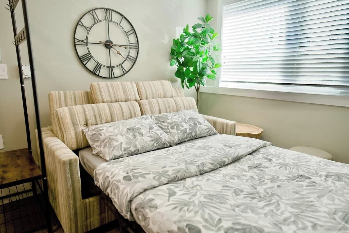 A bed with a floral bedspread, a wall clock, a plant, and a window with blinds.