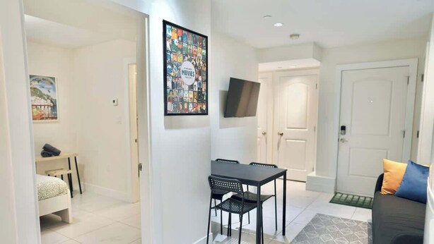 Una sala de estar moderna con un sofá, una mesa de comedor con sillas, un televisor montado en la pared y una puerta que da a otra habitación.