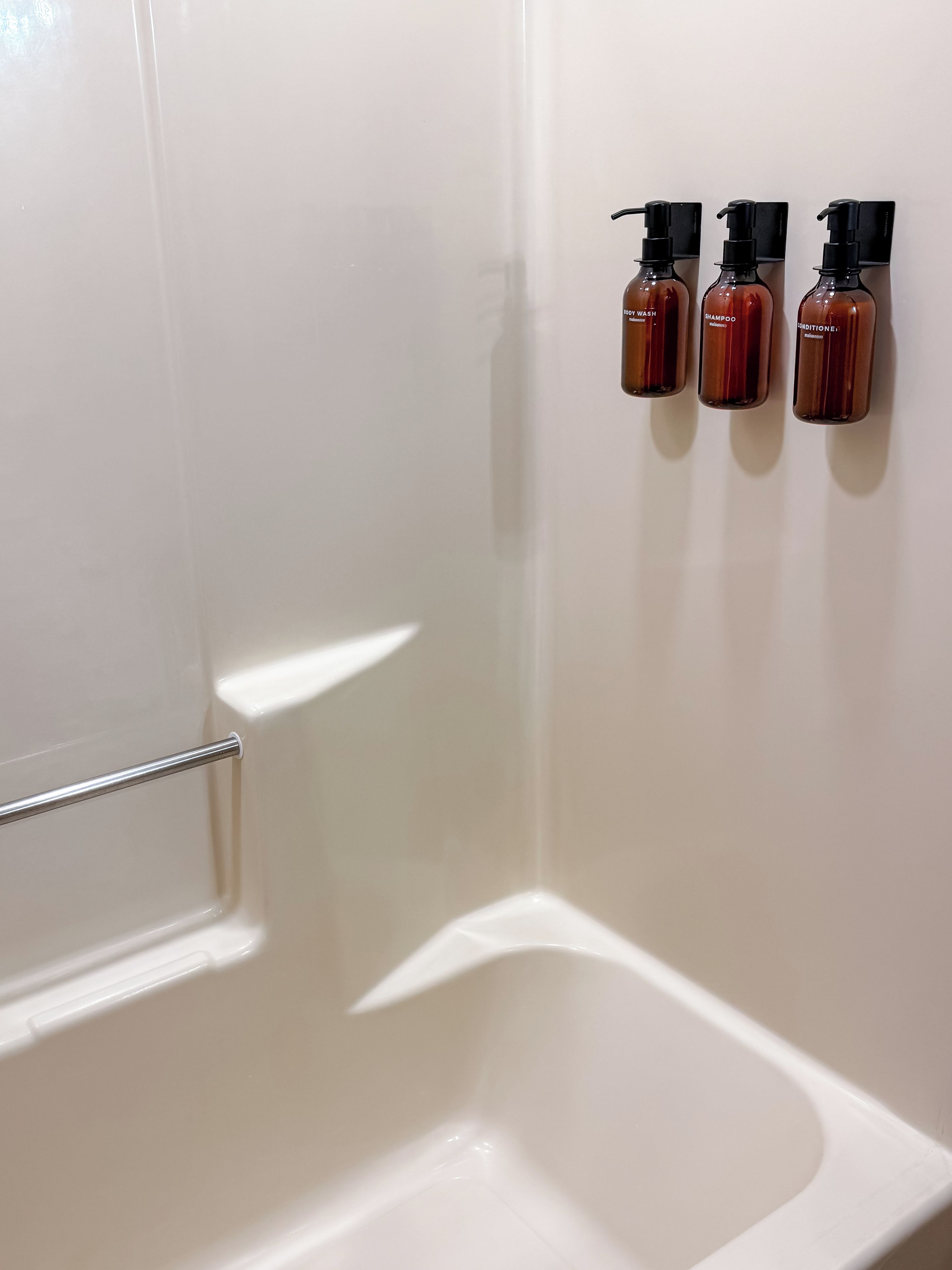 A white bathtub with a handrail and three bottles of shampoo and conditioner on the wall.