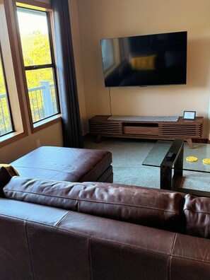 Una sala de estar con un sofá de cuero, un televisor de pantalla plana montado en la pared, una mesa de centro y un ventanal con vista a árboles.