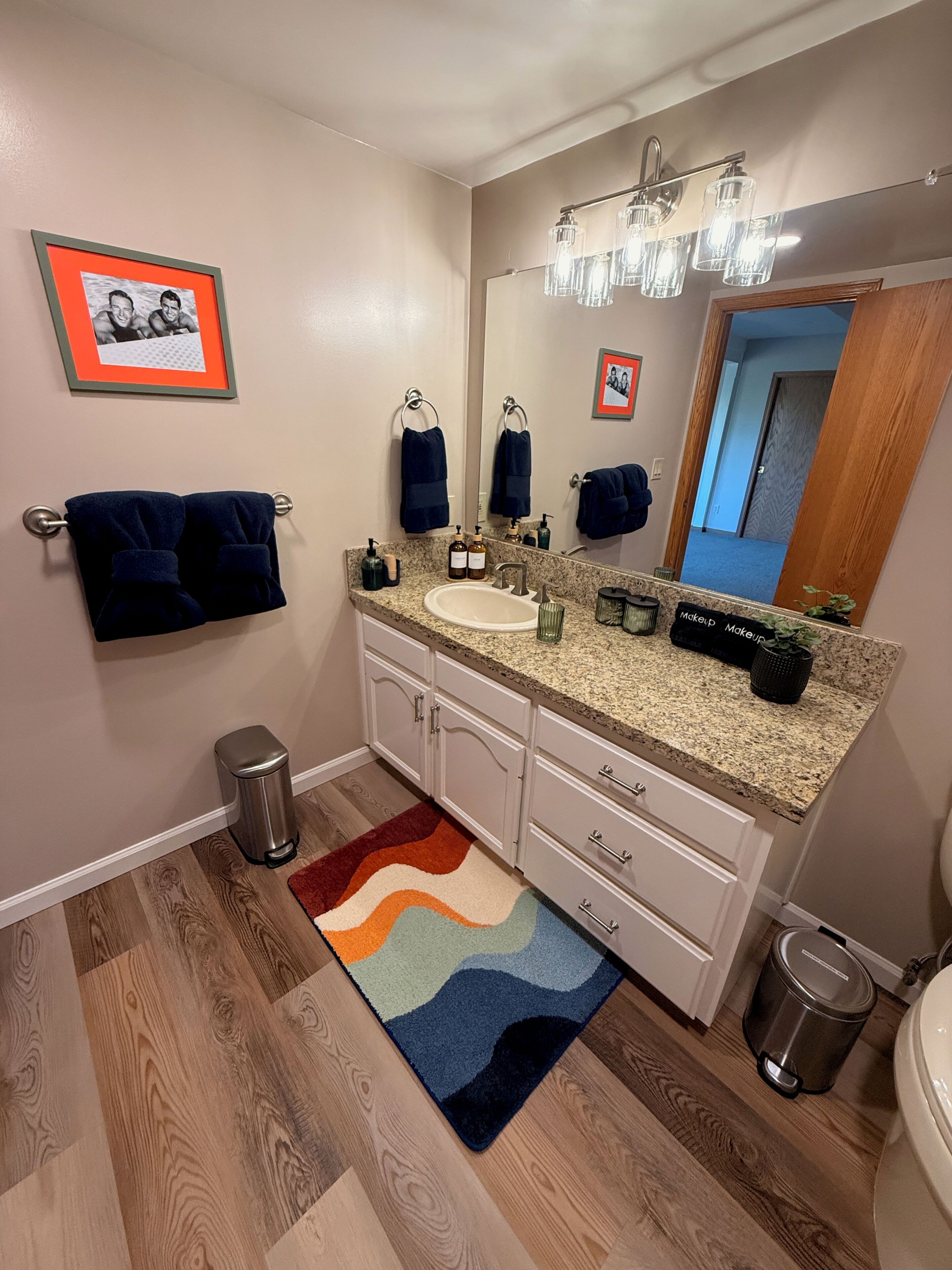 A bathroom with a granite countertop sink, a mirror with lights, a trash can, and a colorful rug.
