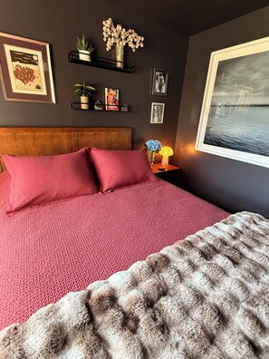 A bedroom with a large bed, a headboard, a nightstand with a lamp, and wall decorations including framed pictures and a vase with flowers.